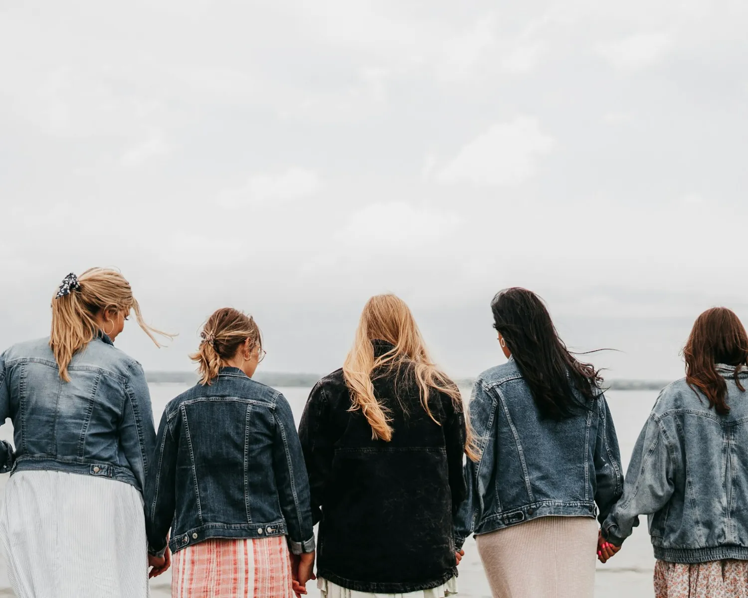 Vijf vrouwen staan naast elkaar aan het water en houden elkaars hand vast, van achteren gezien